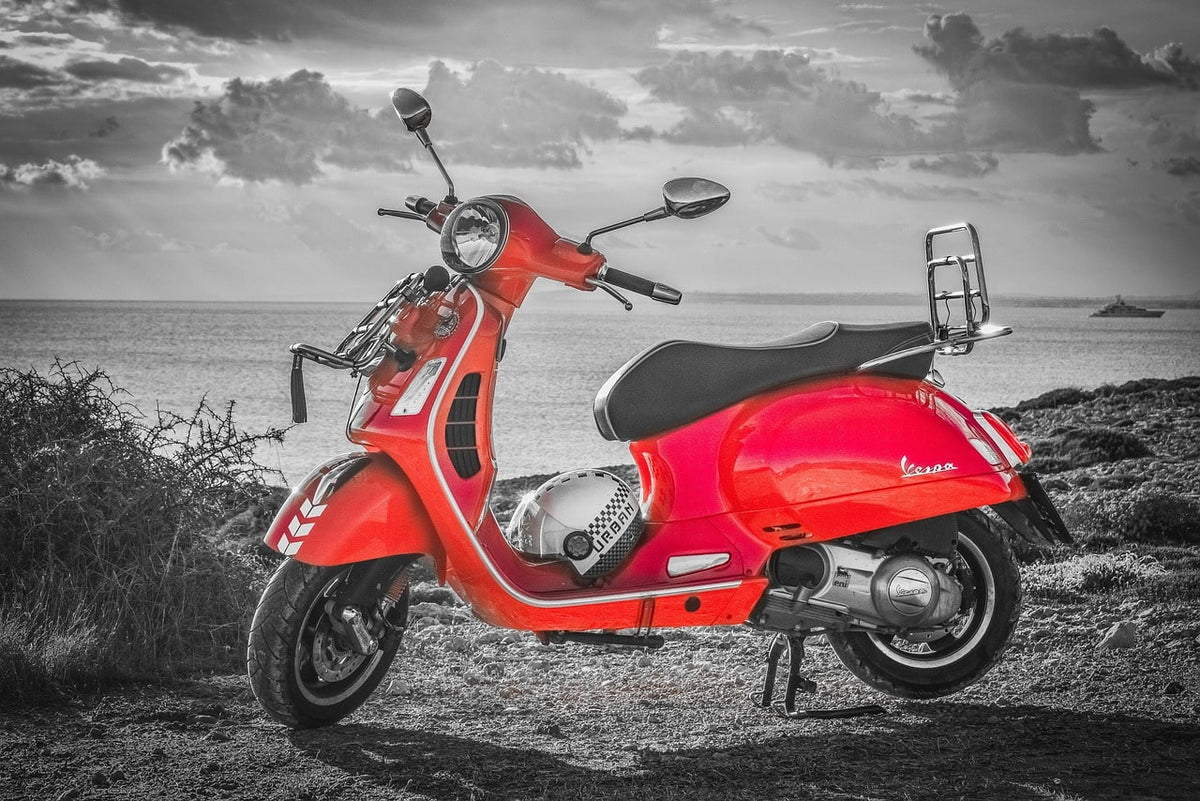 Scooter Vespa vermelha estacionada junto ao mar, em fotografia a preto e branco